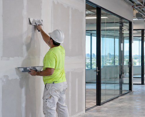 Commercial drywall finisher coating office walls beside glass partitions