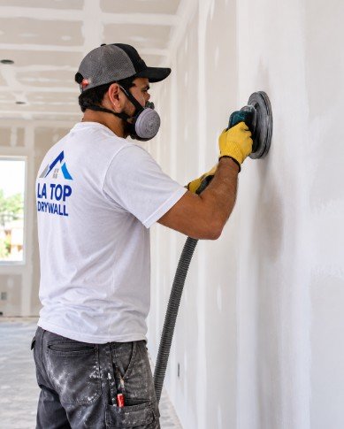 Drywall finisher sanding a wall with a power sander and respirator